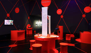 A red table with three seats, each with a roll of coloring pages in front of them. It is an art exhibit at Deutsches Hygiene Museum Dresden