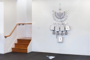 A photo of a room. On a wall next to a staircase hangs a sculpture made of wood, paper, and electronics. It looks like half diagram, half machine.